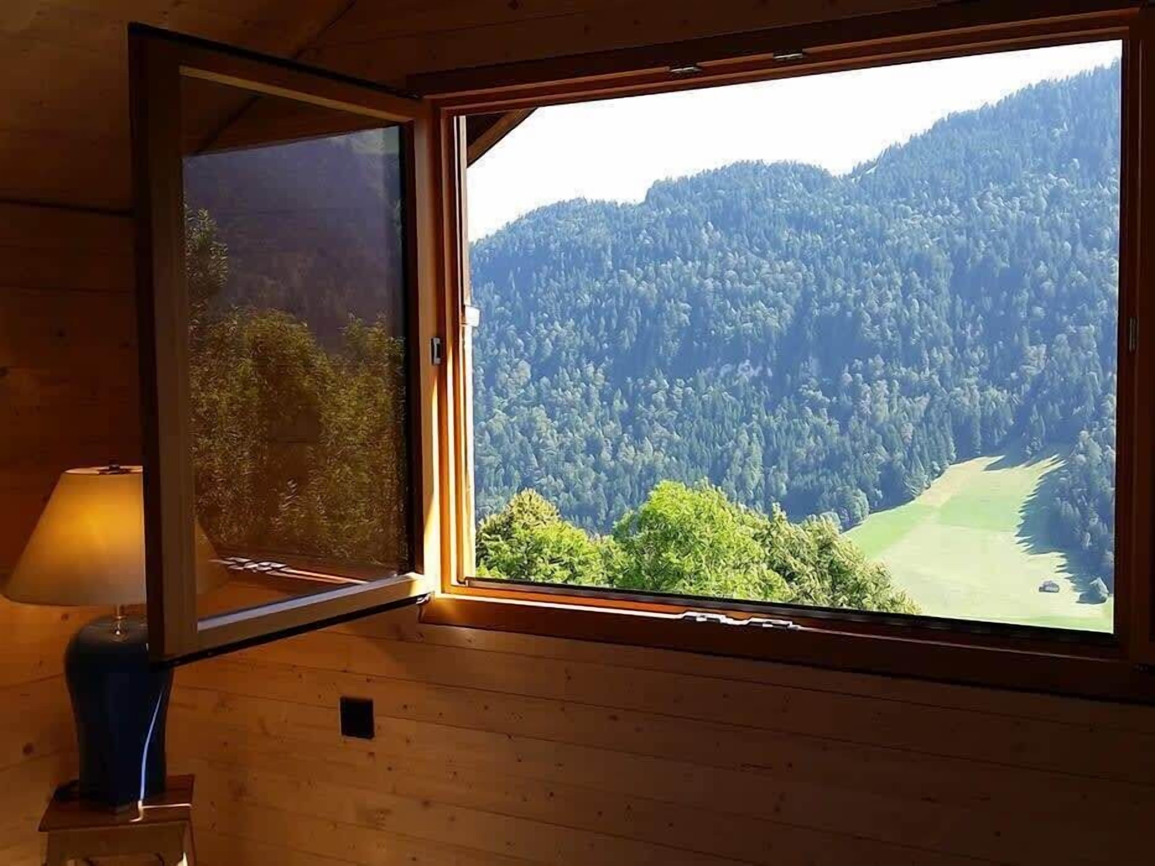 Une pièce au décor intérieur en bois, une fenêtre donnant sur une montagne boisée et une lampe posée sur une table.
