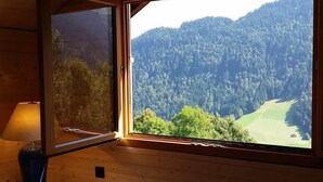A room with a wooden interior, a window with a view of a forested mountain, and a lamp on a table. - Heidi Chalet - Panoramic View - Modern Part (Rossinière)