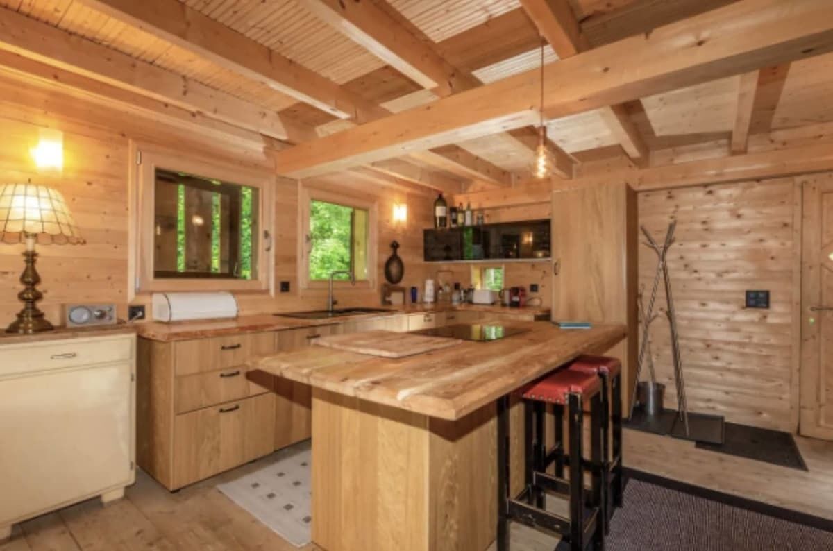Interior de una cabaña de madera con una isla de cocina, taburetes de bar, una ventana y una puerta.