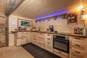 A kitchen with wooden cabinets, a built-in oven, a coffee maker, and a shelf with bottles. - Heidi Chalet Alps - Unique Experience - Authentic Part (Rossinière)
