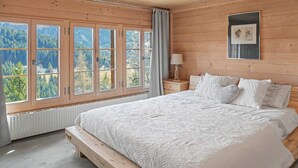 A bedroom with a large bed, a nightstand, a window with a view of trees, and a framed picture on the wall. - Paradise View - 5 BR Gstaad Paradise View Chalet with Outdoor Jacuzzi (Rougemont)