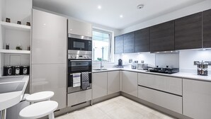 A modern kitchen with white cabinetry, a built-in oven, a microwave, and a coffee maker. There are two white stools and a window providing natural light. - Trevillion Mansions Chiswick (London)