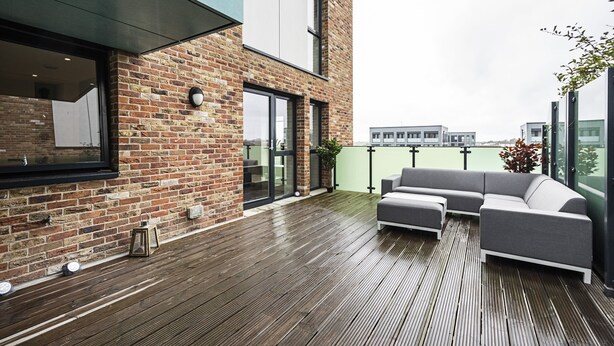 A modern outdoor patio with a wooden deck, a gray sofa, and a brick building with large windows. - Trevillion Mansions Chiswick (London)