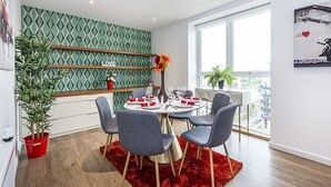 A modern dining area with a round table set for four, surrounded by grey chairs. There are two shelves with decorative items and a potted plant. - Trevillion Mansions Chiswick (London)