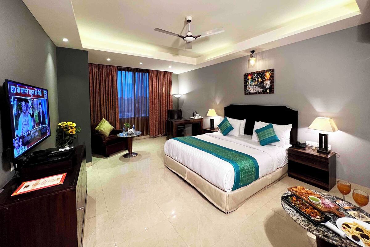 A hotel room with a bed, a TV, a desk, a chair, a table with food, and a ceiling fan.