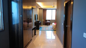 A hallway with a dark wooden wardrobe, a flat-screen TV on a stand, a small table with a chair, and a bed with a red blanket. - The Shoba Estate Gold (New Delhi)