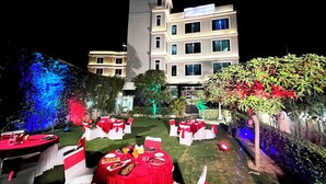 An outdoor dining area with tables set for a meal, surrounded by greenery and a building in the background. - The Shoba Estate Gold (New Delhi)