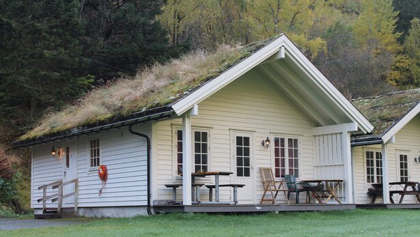 A two-story house with a sloped roof covered in vegetation, a front porch with outdoor seating, and a small front yard. - Hjelledalen Hyttesenter (Stryn)