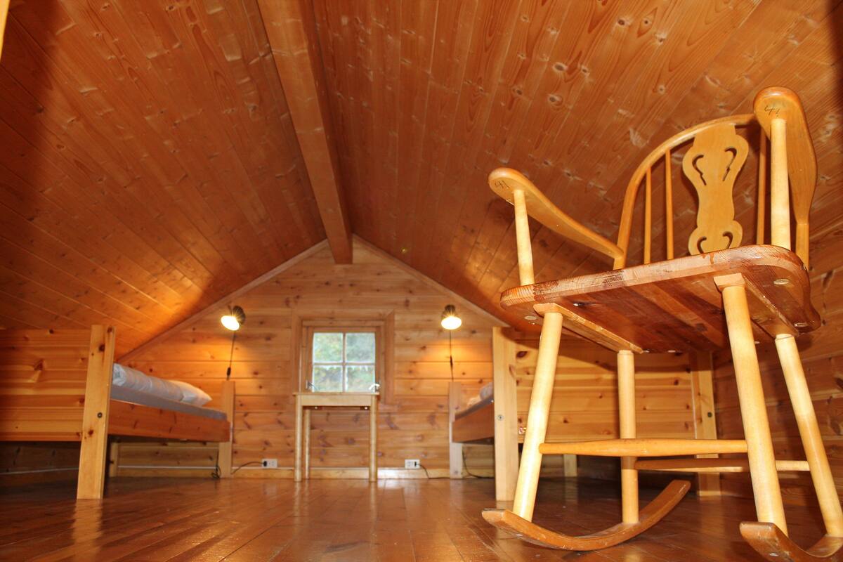 A wooden cabin with a sloped ceiling, a rocking chair, a window, and a bed.