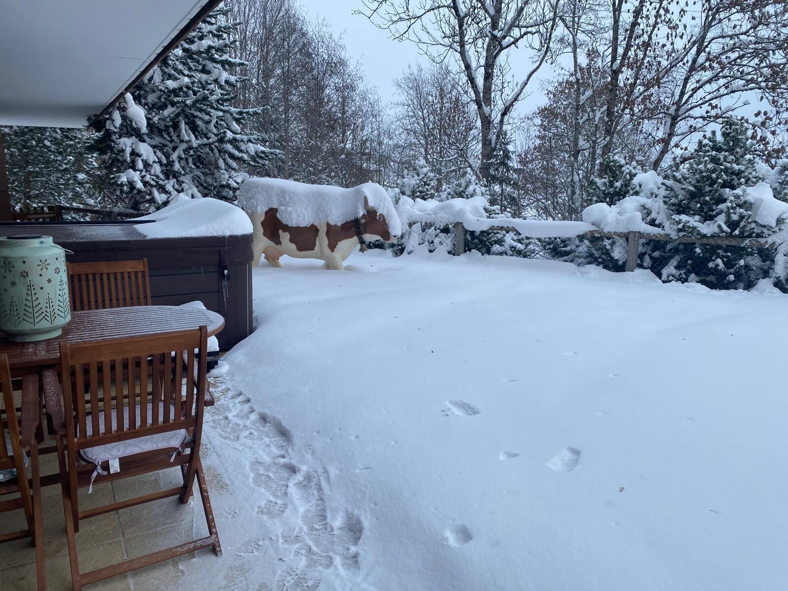 Une terrasse enneigée avec une table et des chaises en bois, un pot en céramique décoratif et une statue de vache recouverte de neige.