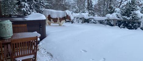 Une terrasse enneigée avec une table et des chaises en bois, un pot en céramique décoratif et une statue de vache recouverte de neige.