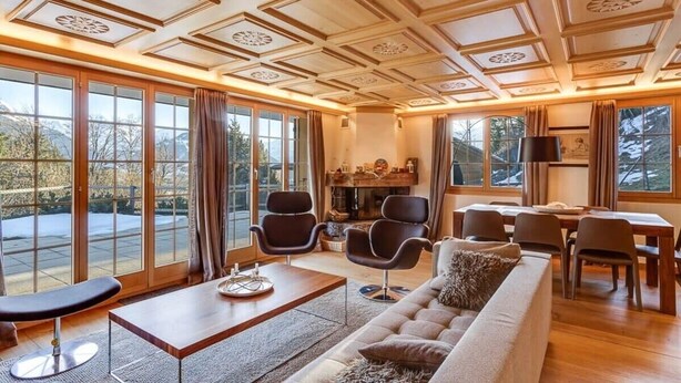 A spacious living room with a large sofa, a coffee table, and a fireplace. There are multiple chairs and a dining table. The room has large windows with curtains, and the ceiling features decorative panels.