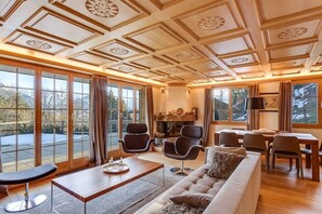Ein geräumiges Wohnzimmer mit einer großen Couch, einem Couchtisch und einem Kamin. Mehrere Stühle und ein Esstisch sind ebenfalls vorhanden. Große Fenster mit Vorhängen und dekorative Paneele an der Decke runden den Raum ab.