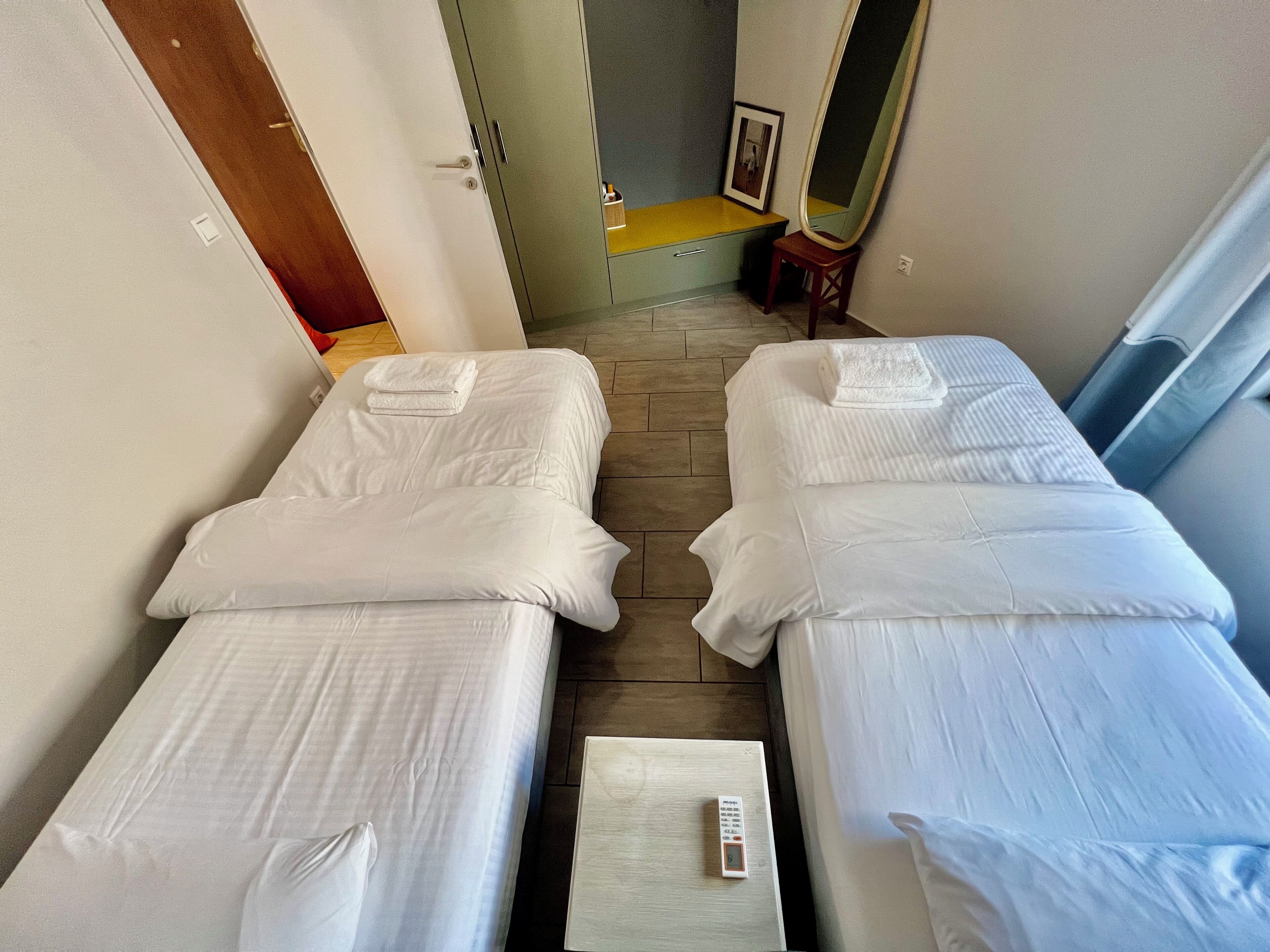 A hotel room with two single beds, a small table with a remote control, a mirror, and a framed photo on the wall.