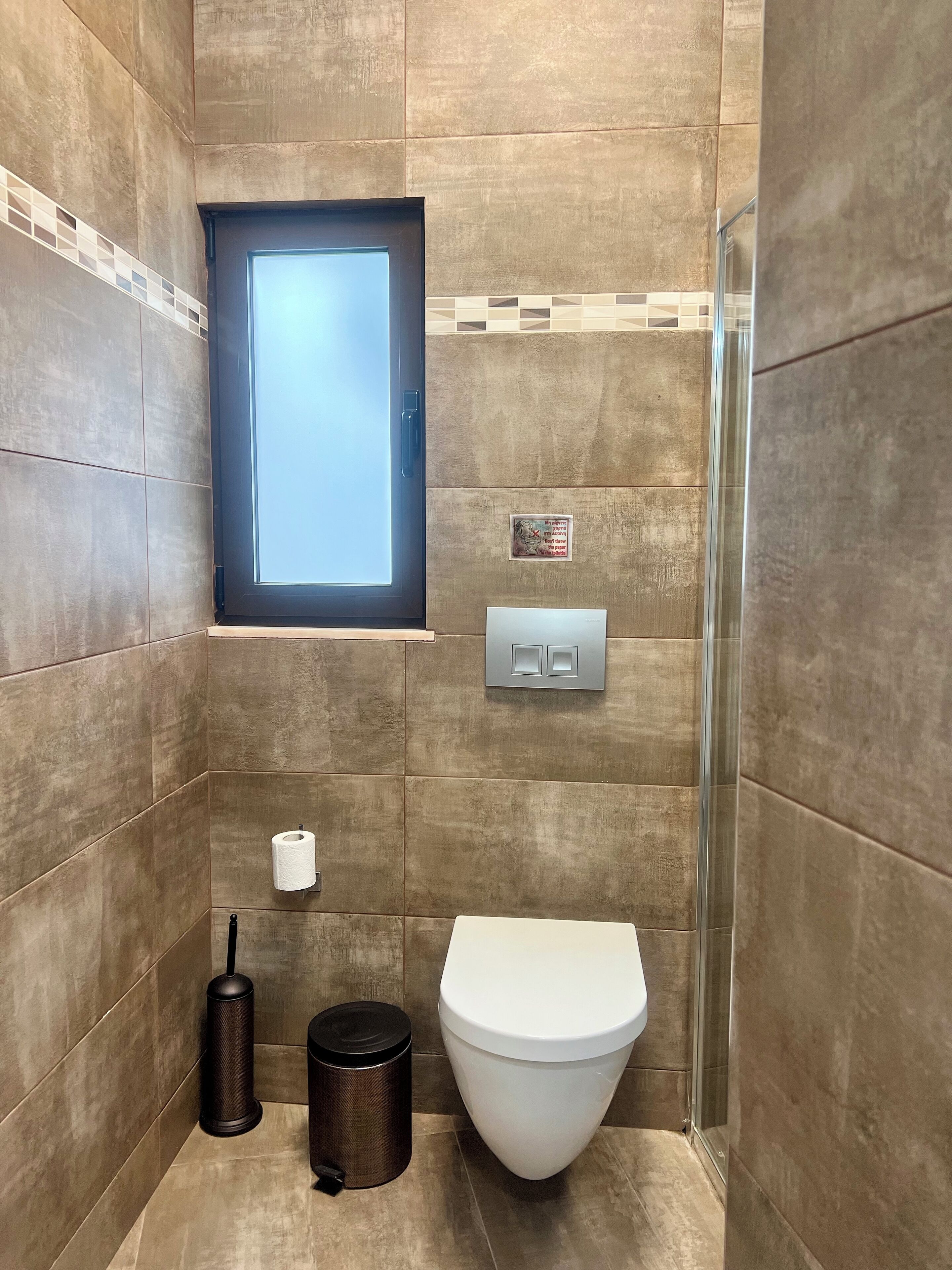 Een badkamer met een toilet, een prullenbak, een handdroger, een raam en een lichtschakelaar.