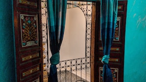 Ein Zimmer mit Fenster, dessen dekorative Eisenverzierungen und zurückgebundene Vorhänge zu sehen sind, ein Stuhl vor dem Fenster und ein gefliestes, gemustertes Parkett.