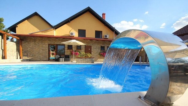 A swimming pool with a water feature, a brick building with a yellow facade, and a clear blue sky. - Guest House Vila Alexandar (Palic)