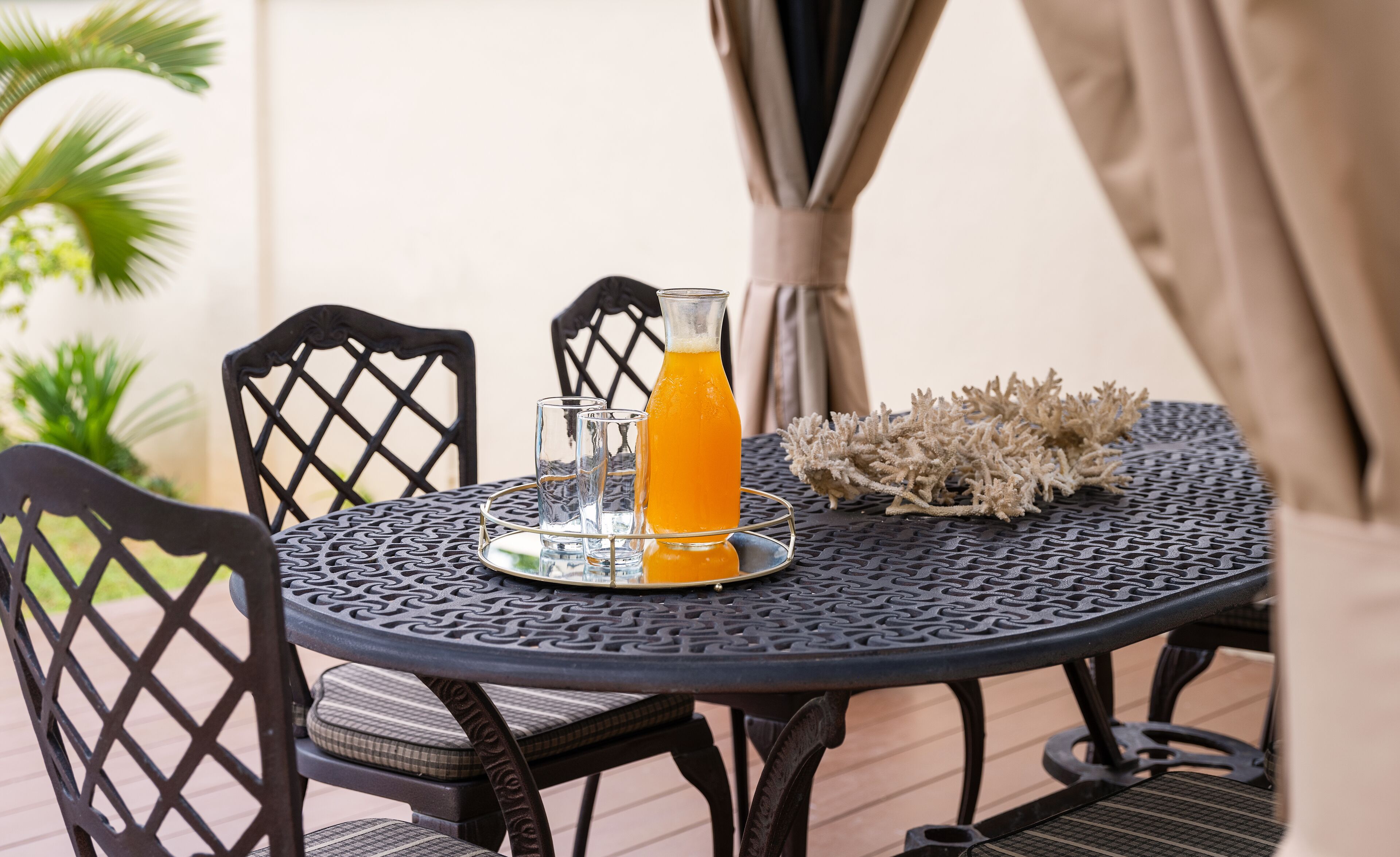A round metal table with a patterned top, surrounded by four chairs with a lattice back design. A clear glass pitcher with a yellow liquid and two empty glasses are on the table.