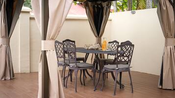 A covered outdoor dining area with a round table and four chairs, surrounded by beige curtains.