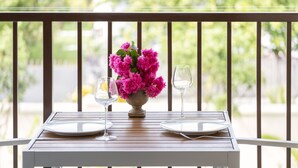 Una mesa con dos platos blancos, un florero de flores rosadas y dos copas de vino sobre una mesa de madera en un balcón.