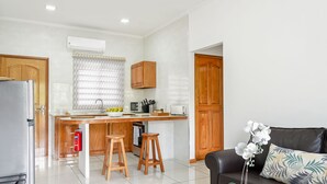 Una cocina moderna con una isla central, armarios de madera y un microondas integrado. Hay un refrigerador a la izquierda, un fregadero con una ventana encima y un extintor debajo del mostrador. En la zona de estar hay un sofá de cuero negro con un cojín decorativo.