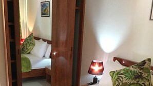 Una cama bien hecha con colcha verde, almohadas blancas y una almohada estampada. Se ve un armario de madera con espejo, una lámpara de noche y un cuadro en la pared.