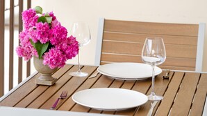 Una mesa de comedor con dos platos blancos, dos copas de vino, un florero con flores rosadas y un tenedor y cuchillo sobre una mesa de madera, con una silla blanca al fondo.