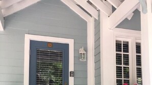 Una puerta azul con una ventana, un porche blanco con un farol, y una maceta con flores.