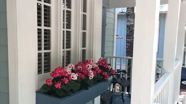 Un porche cubierto con una jardinera integrada llena de flores, una silla y vista al jardín trasero.