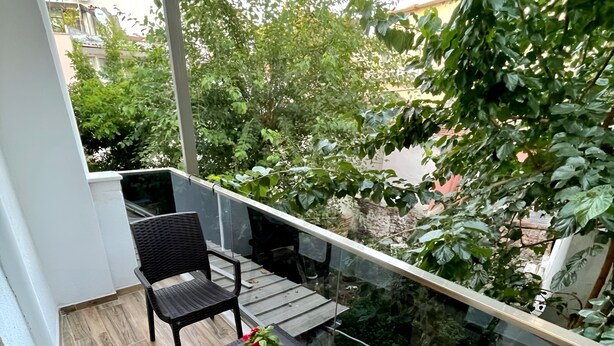 Un balcón con mobiliario de mimbre, una pequeña mesa con una planta y vistas a zonas verdes y edificios.