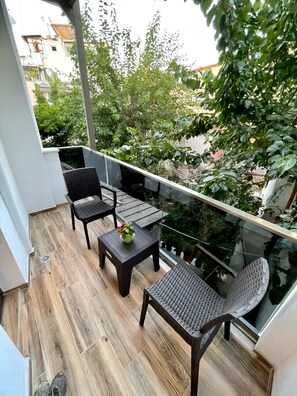 Een balkon met rieten meubels, een tafeltje met een plant en uitzicht op groen en gebouwen.