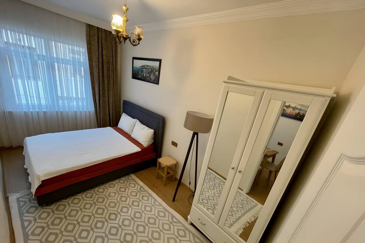 Quarto de hotel com cama, guarda-roupa, candeeiro de chão e janela com cortinas.