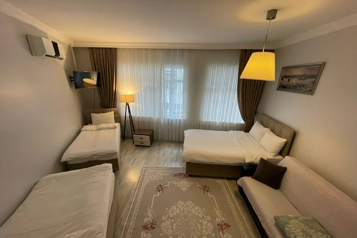 Quarto de hotel com duas camas de solteiro, uma cama de casal, um sofá, um abajur, uma mesa lateral e um tapete decorativo.