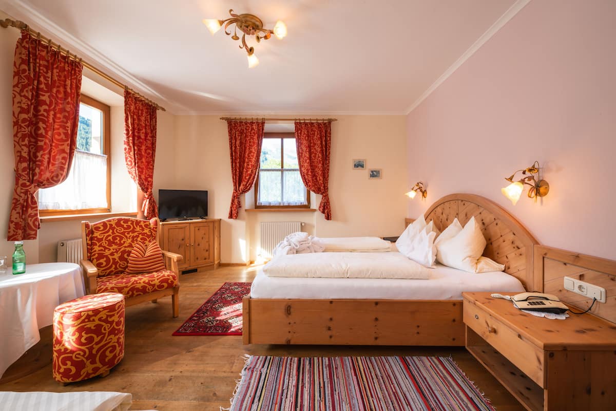 A bedroom with a wooden bed, a chair, a table, a TV, and a window with curtains.