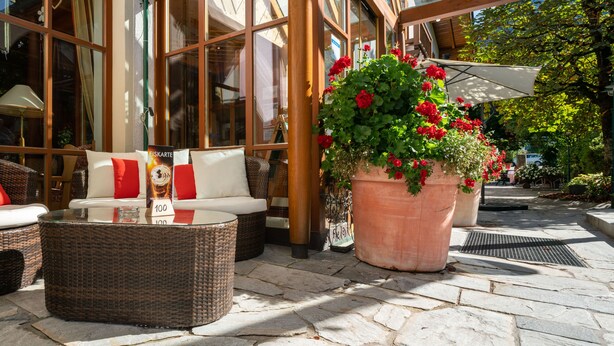 An outdoor seating area with wicker furniture, a glass-top table, and a large potted plant with red flowers. - Hotel Krimmlerfälle (Krimml)