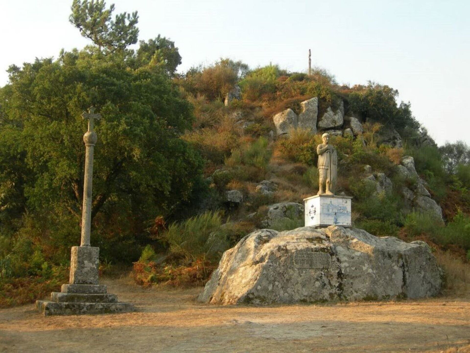 Une statue sur un rocher, avec une plaque commémorative, un pilier de pierre et, au loin, une colline parsemée d’arbres.
