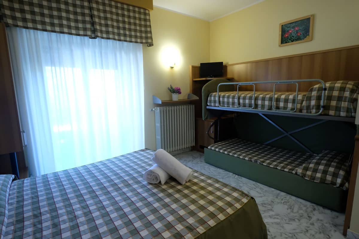A hotel room with a bed, a bunk bed, a radiator, a television, and a window with curtains.
