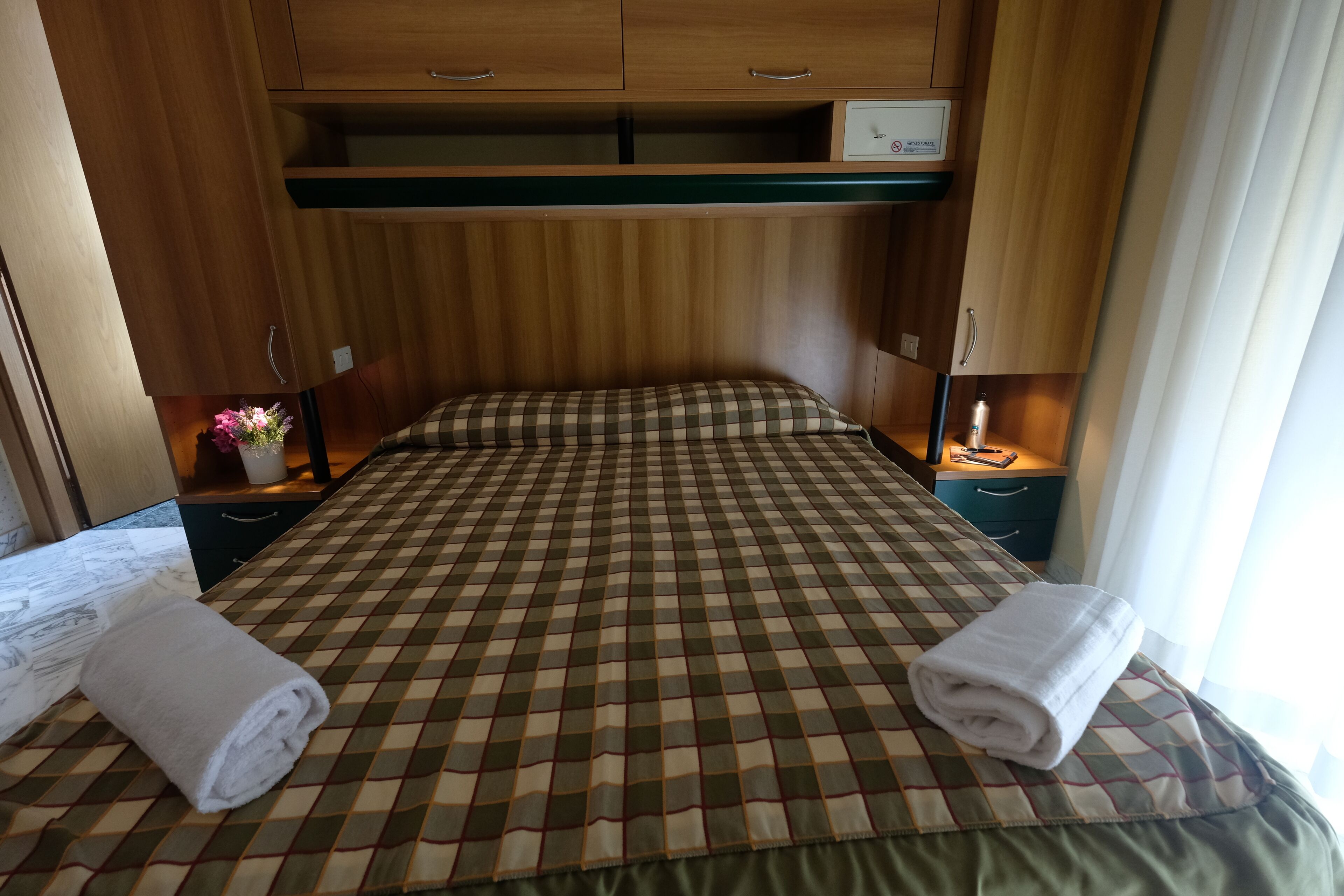 A hotel room with a checkered bedspread, two bedside tables, a nightstand, a small cabinet, and a window with curtains.