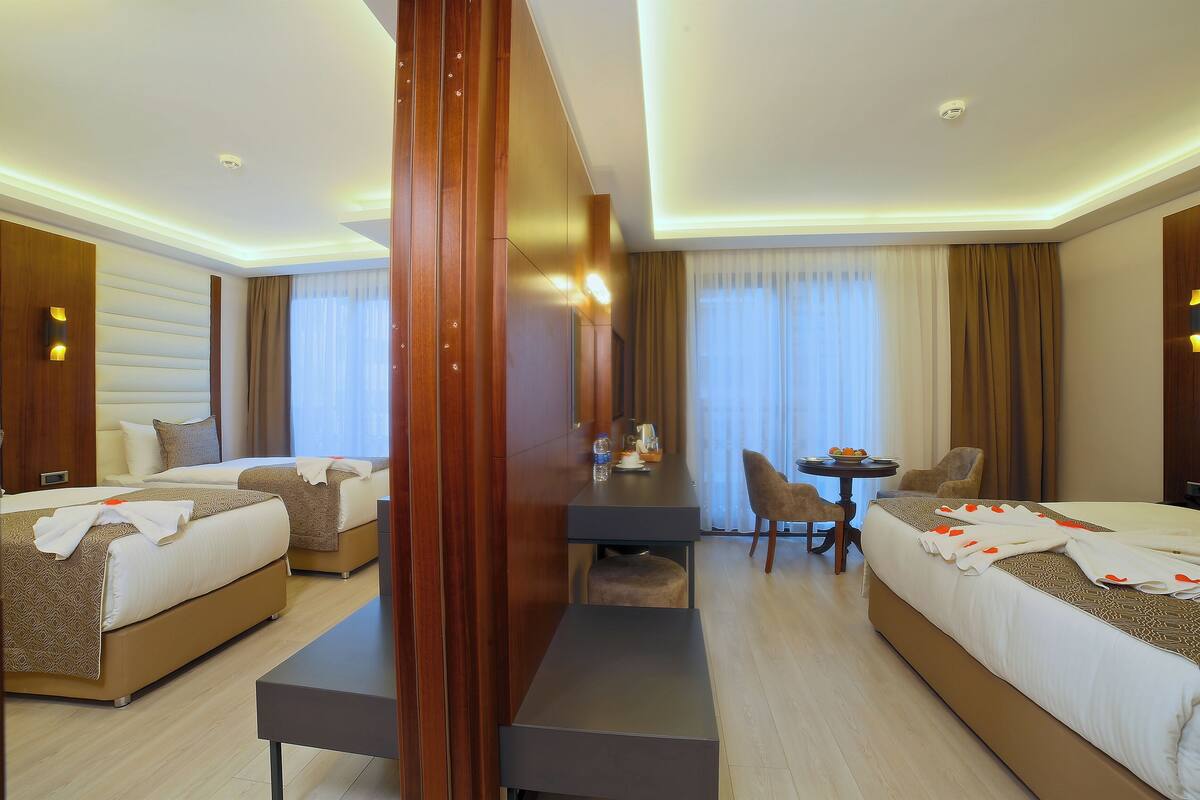 A hotel room with two beds, a desk, a chair, and a small table with a bottle and glasses.
