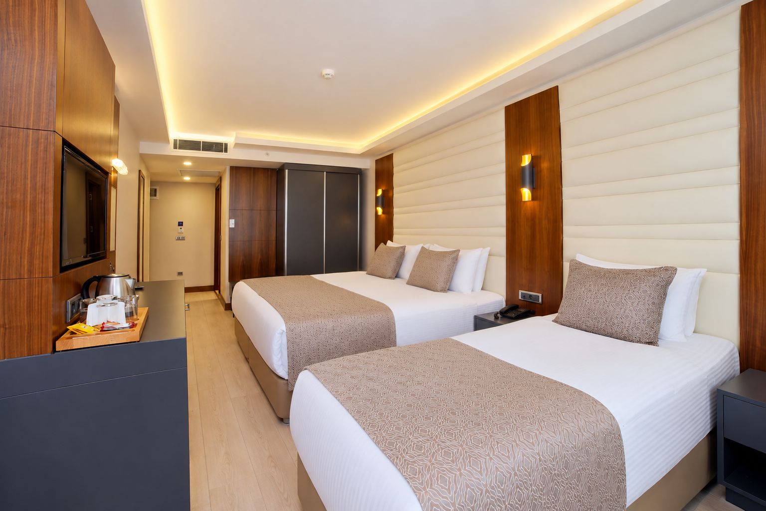 A hotel room with two beds, a desk with a kettle and glasses, and a TV on the wall.