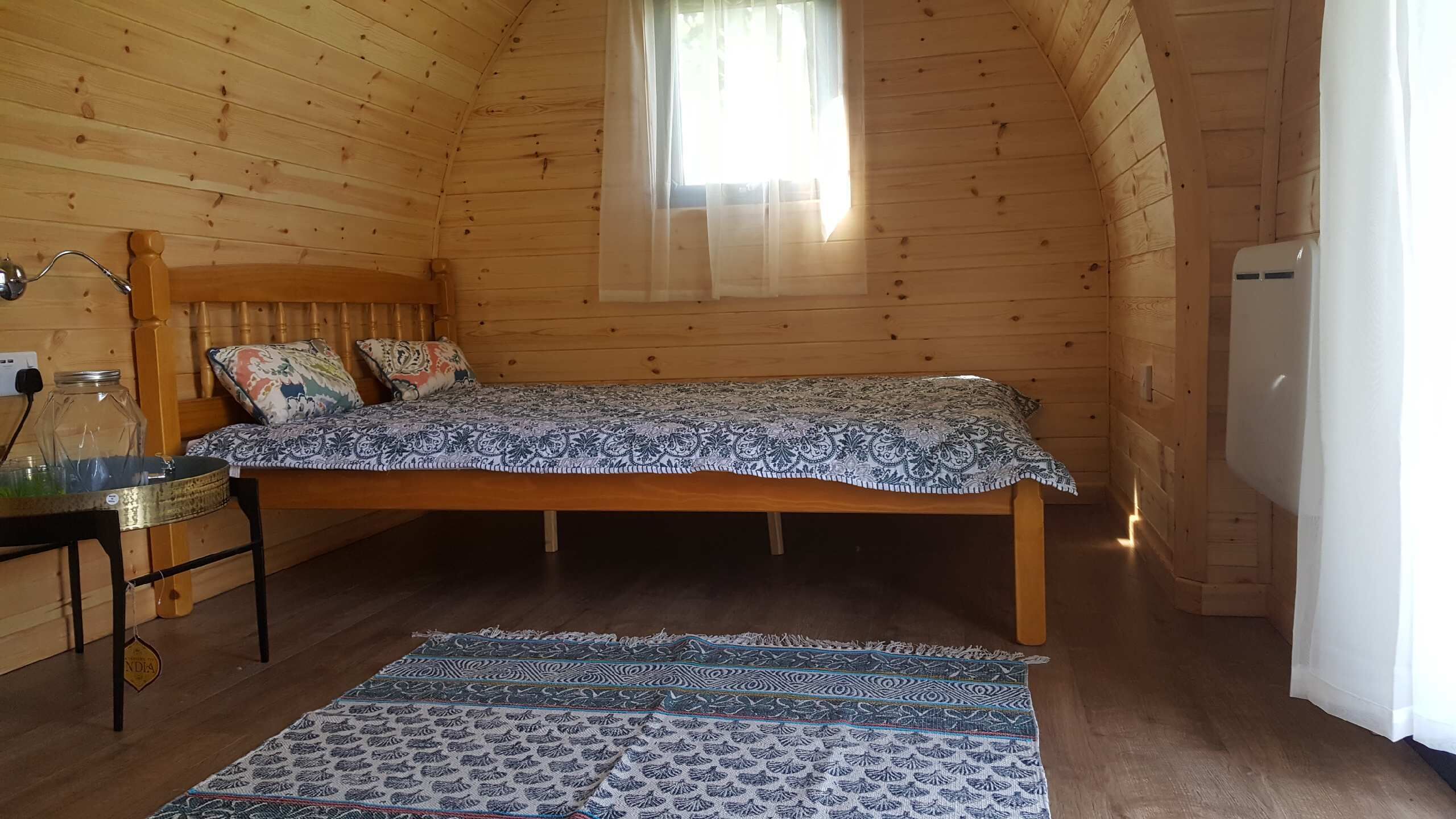 A cozy wooden cabin room with a bed, a small table with a glass jar, and a patterned rug.