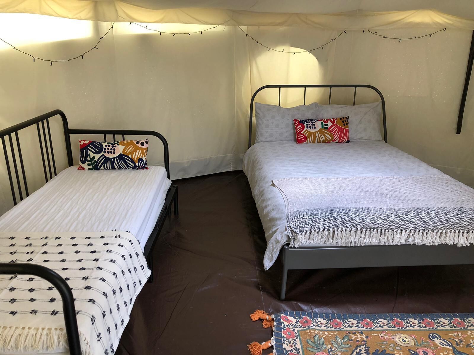 Two single beds with metal frames, each with a patterned bedspread and a decorative pillow. The room has a brown floor and a colorful rug.