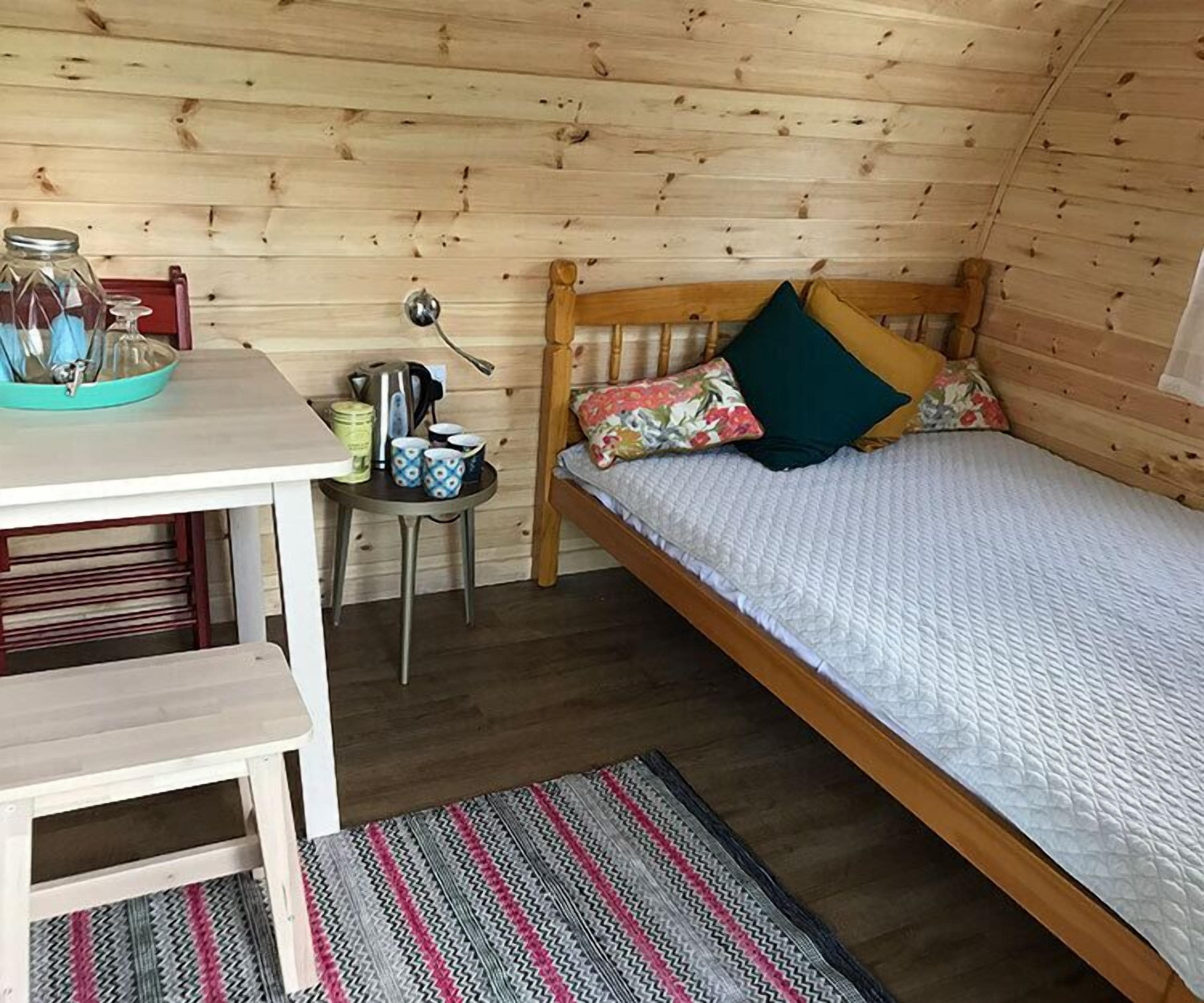 Acogedora habitación tipo cabaña de madera, con cama, una pequeña mesa con utensilios de cocina y un tapete a rayas.