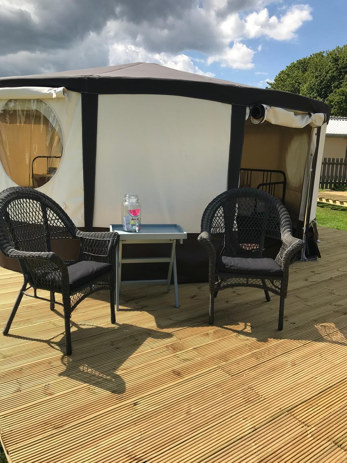Un espace extérieur abrité avec deux fauteuils en osier et une table, une petite table avec un pichet d’eau, et une tente en arrière-plan.