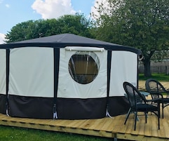 Una carpa grande, blanca y negra con un ventanal circular, ubicada sobre una plataforma de madera con dos sillas negras.