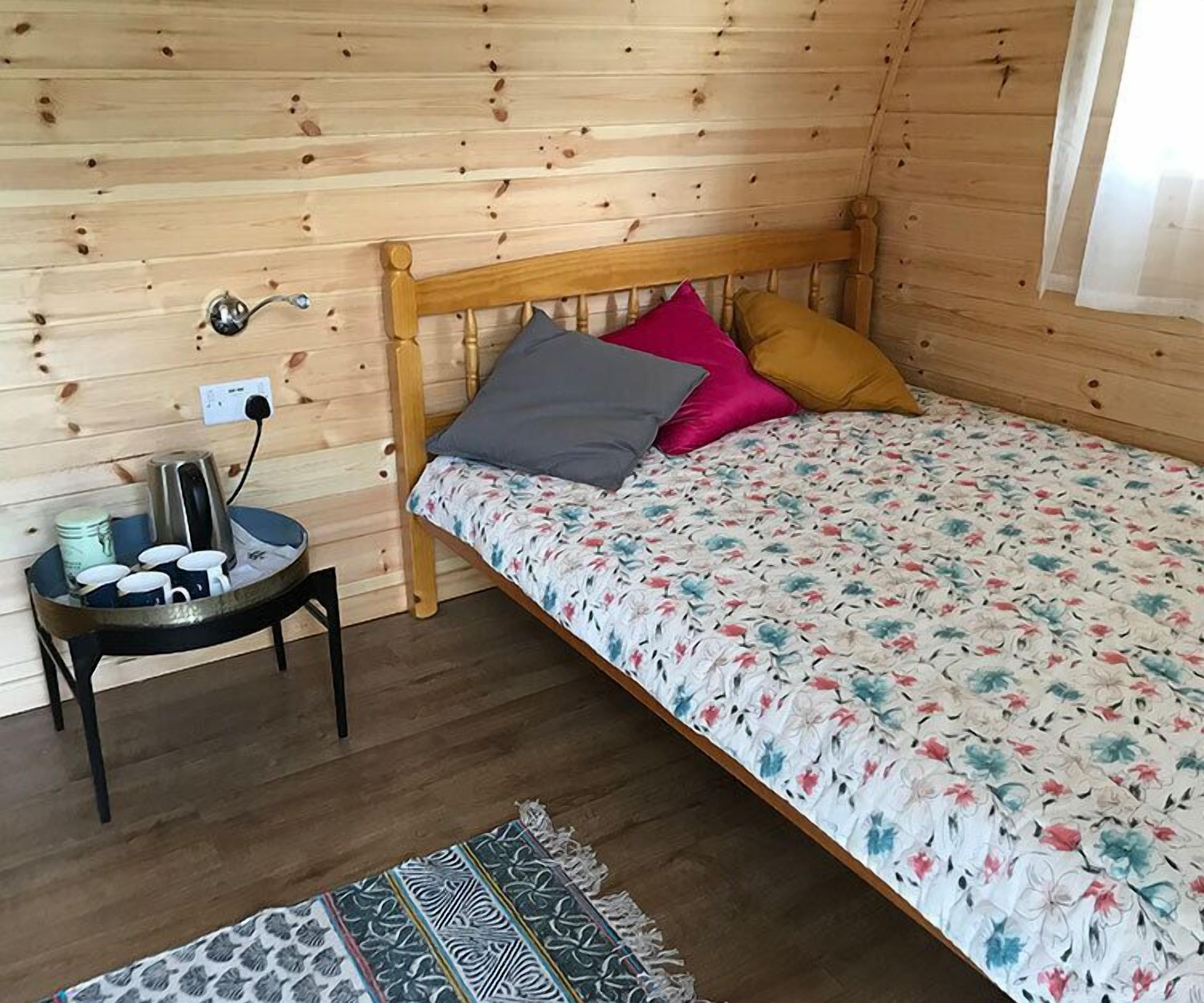 A cozy wooden cabin room with a bed, a small table with a kettle and cups, and a patterned rug.