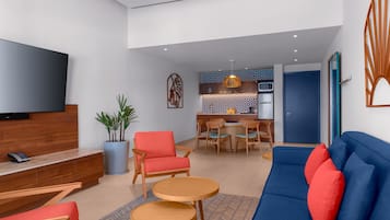 Una sala de estar moderna con un sofá, sillones y un televisor. Se observa un comedor con una mesa y sillas al fondo.