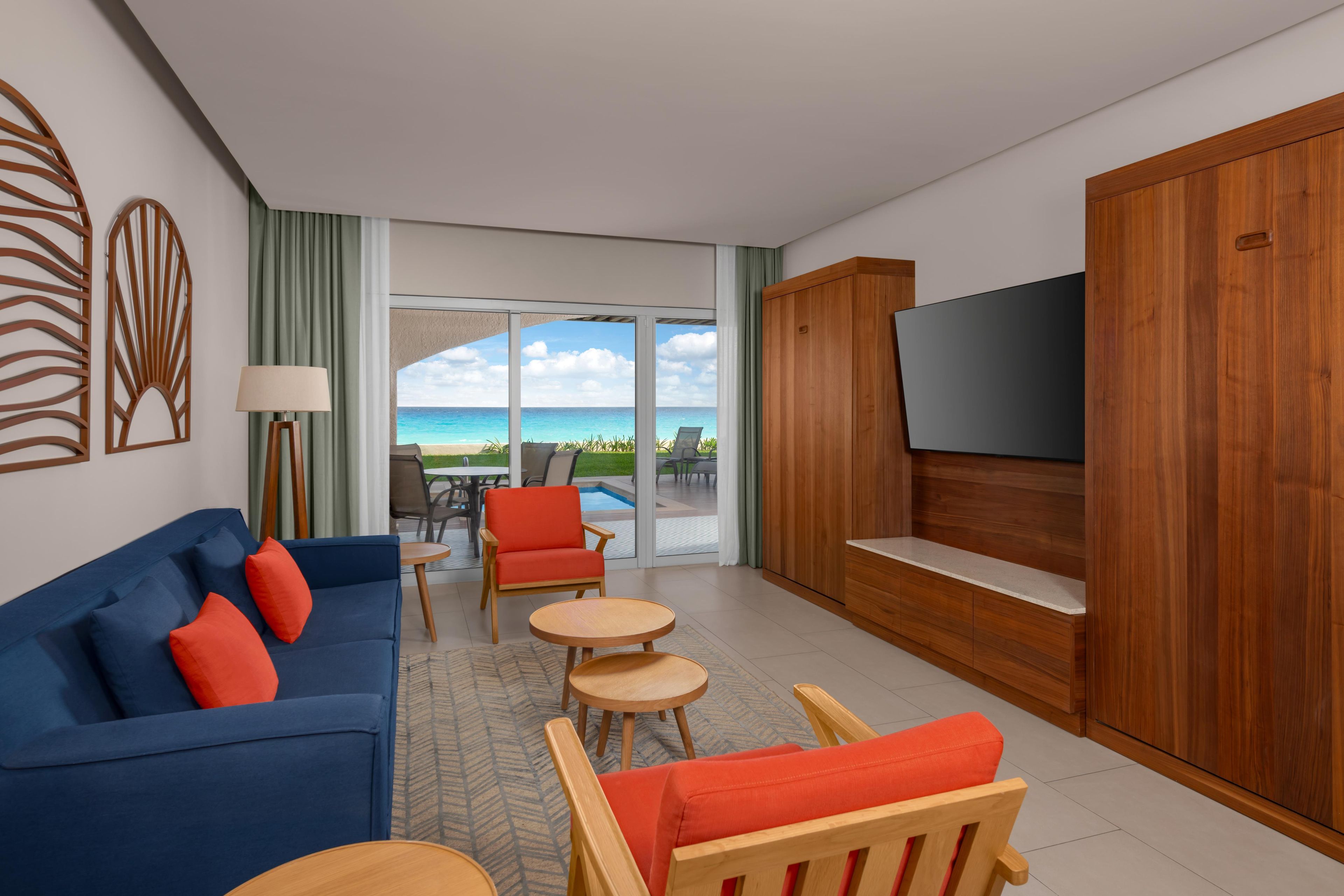 A modern living room with a blue sofa, red cushions, a wooden TV stand, and a view of the ocean through large glass doors.