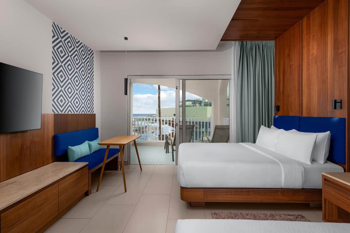 A modern hotel room with a large bed, a blue sofa, a wooden bench, and a balcony with a view.