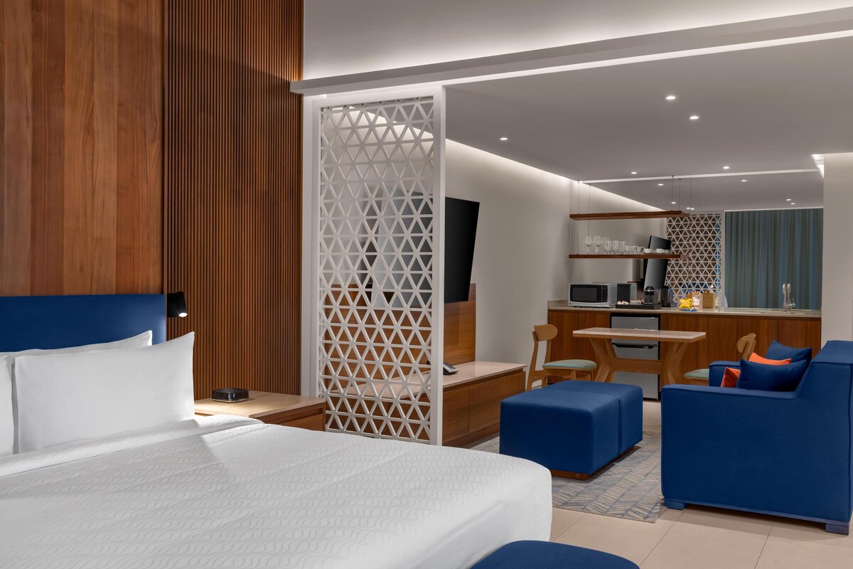 A modern hotel room with a large bed, a desk, a kitchenette, and a seating area with blue sofas.
